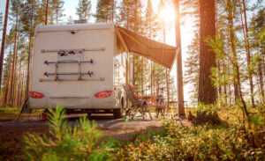 Motor home parked at campsite in woods. Woman lounging in camp chair in sunlight. 