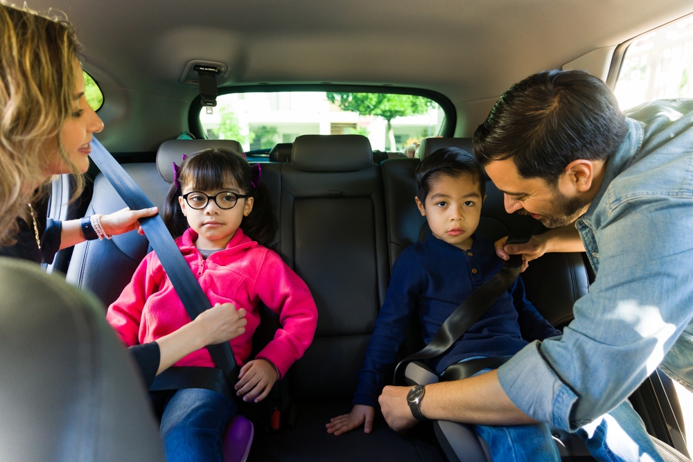 Smiling caucasian parents putting their children into the back seat with a booster seat and seat belt while going on a car trip