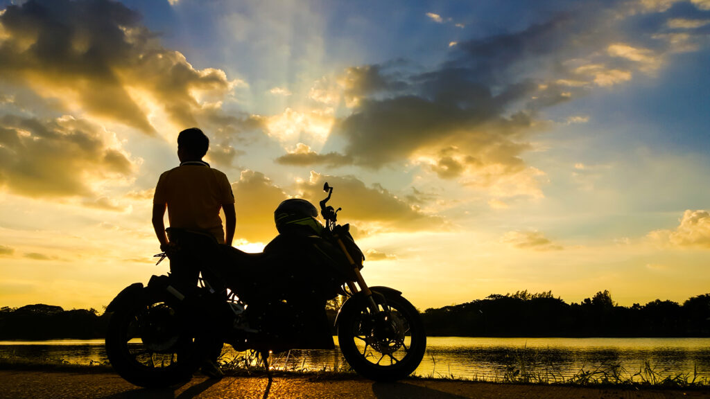 Motorcyclist leaning against bike posing in sunset by lake.